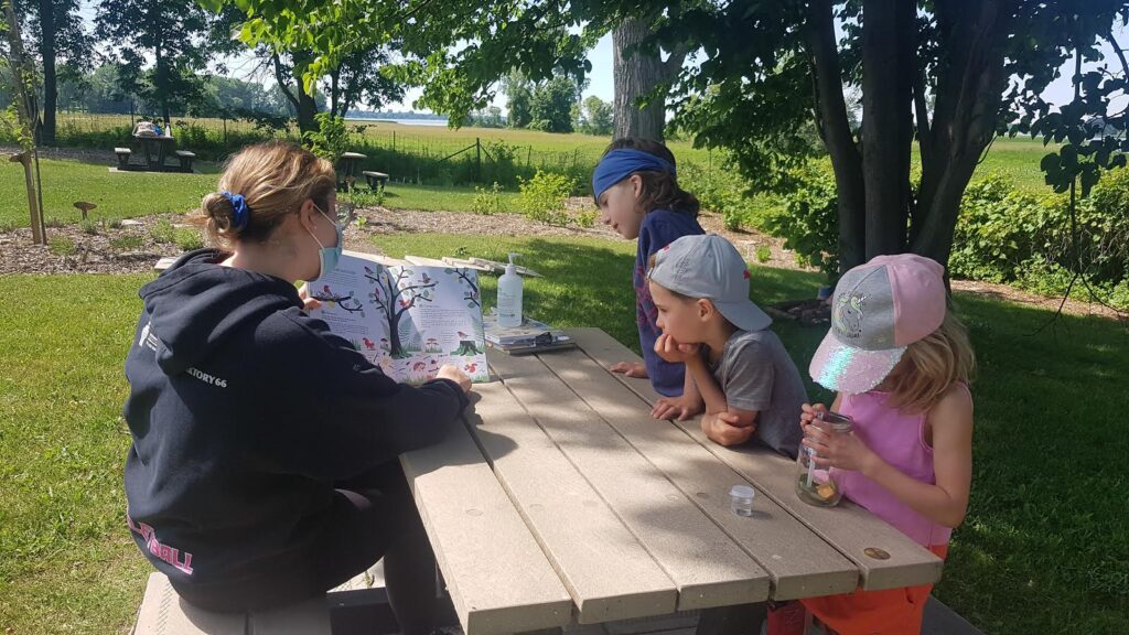 Three children and an adult sit at a picnic table. The adult is showing an illustration of a tree to the kids.