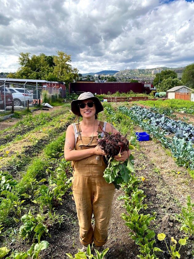 A person in overalls holds a bushel of beets in a garden.