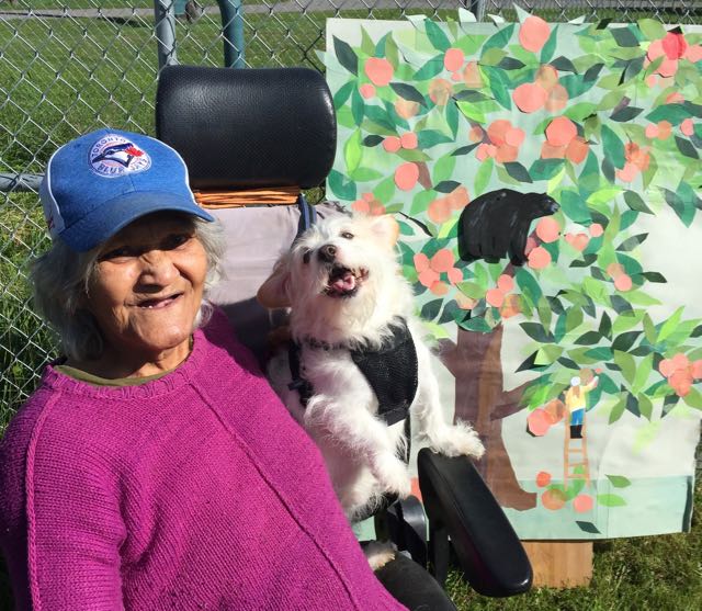 An elderly person in a mobility chair with a small dog smiles in front of a print out of an apple tree and a bear.
